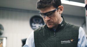 A man wearing safety glasses and a green vest with Continuum Equity Partners embroidered on it is focused on a task. The background includes industrial elements, suggesting hes in a workshop or factory.