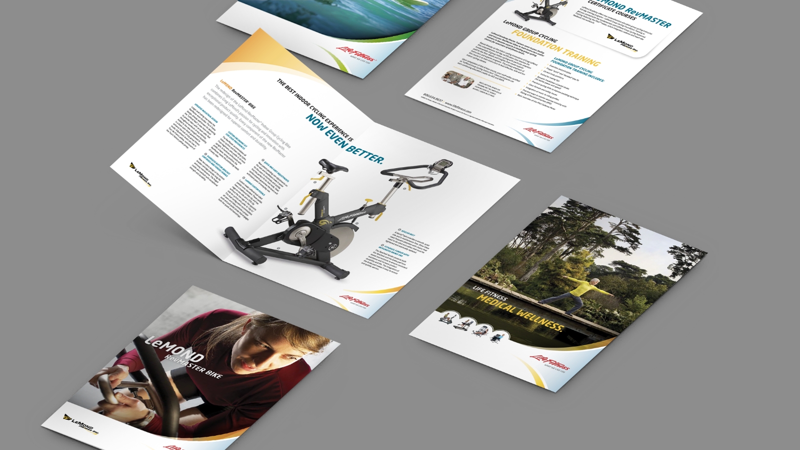 Several fitness brochures are scattered on a gray surface, featuring images of exercise equipment and people working out. The materials showcase product details, benefits, and health tips, with colorful graphics and text in a dynamic layout.