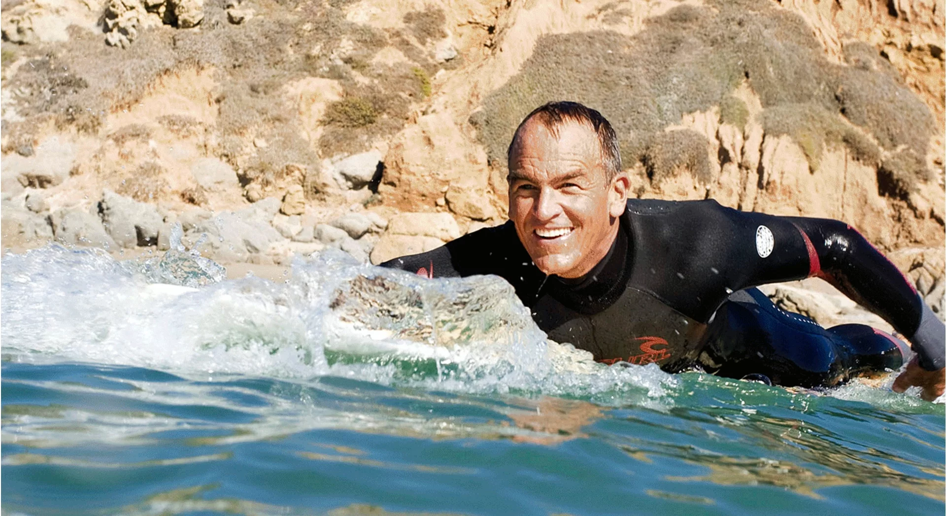 LifeFitness Hero Photo of well-aged man paddling out into water on surfboard, smiling.
