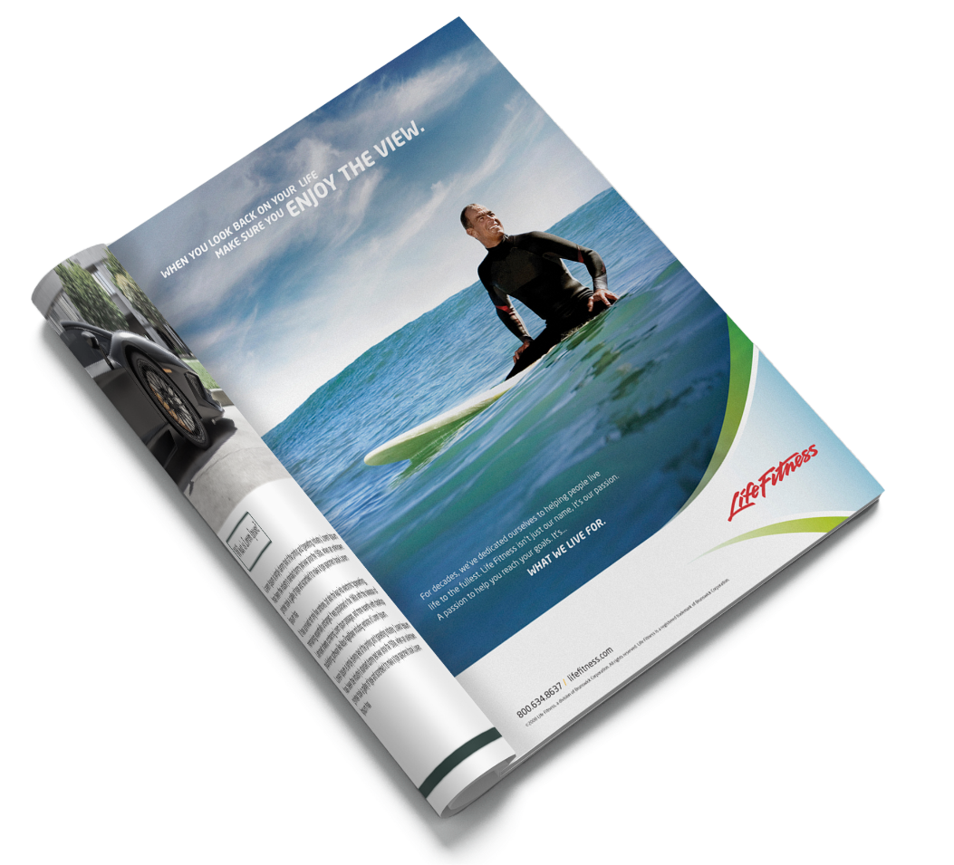 A magazine is open, displaying an advertisement showing a person seated on a stand-up paddleboard, gazing over calm blue water. Text reads Enjoy the view. The Life Fitness logo is visible in the corner.