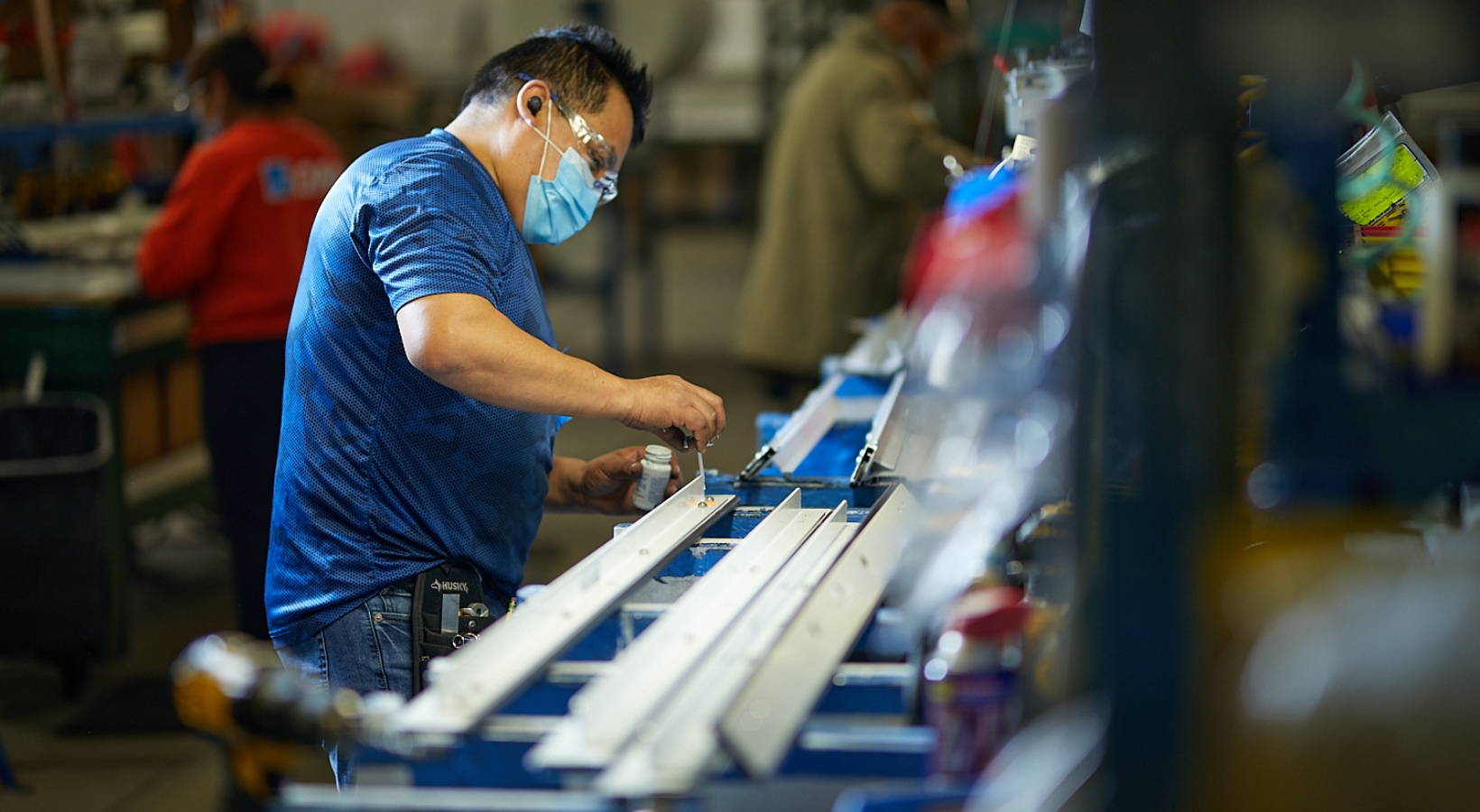 A person wearing a face mask and safety goggles works on a conveyor belt in a factory setting. They are adjusting components on white metal frames, surrounded by various tools and equipment. Other workers are visible in the background.