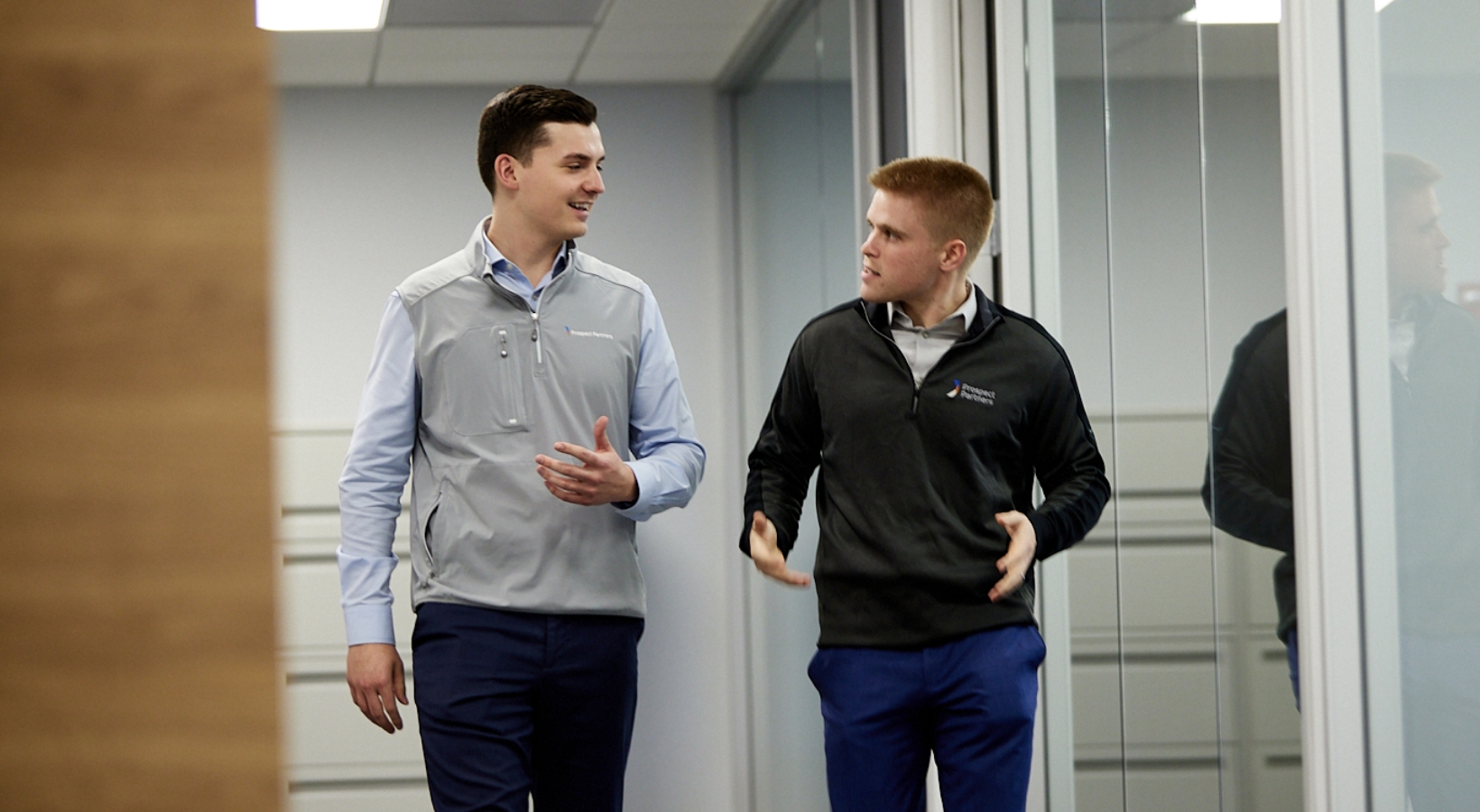 Two men walking and talking in an office hallway. One wears a light gray vest over a white shirt, the other a dark zip-up jacket. They appear engaged in conversation, with lockers and glass doors in the background.