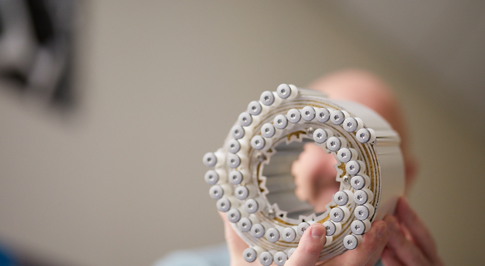 A person holds a spherical object made of stacked AA batteries with white tops, facing the camera. The batteries are arranged in concentric circles, and the background is blurred.