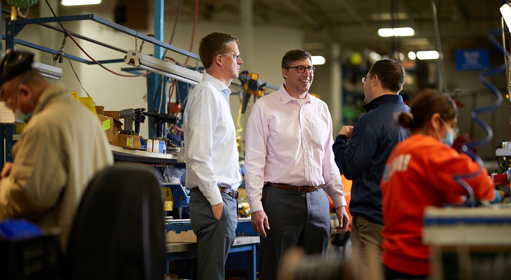 Four people are standing and conversing in a busy workshop setting. Various tools and parts are visible in the background. The focus is on three men smiling and chatting near a workbench, while a woman works nearby.