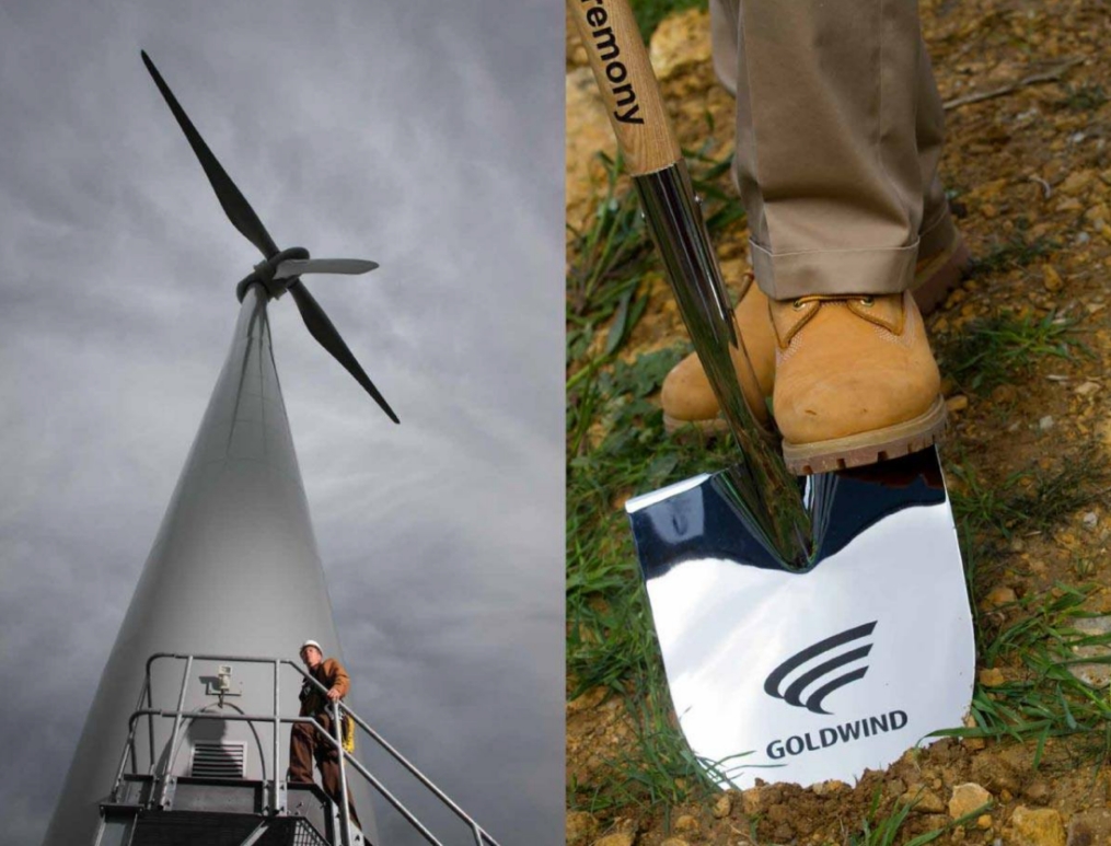 A tall wind turbine viewed from below under a cloudy sky is on the left. On the right, a person wearing tan pants and brown boots stands with a shiny shovel marked Goldwind digging into the earth.