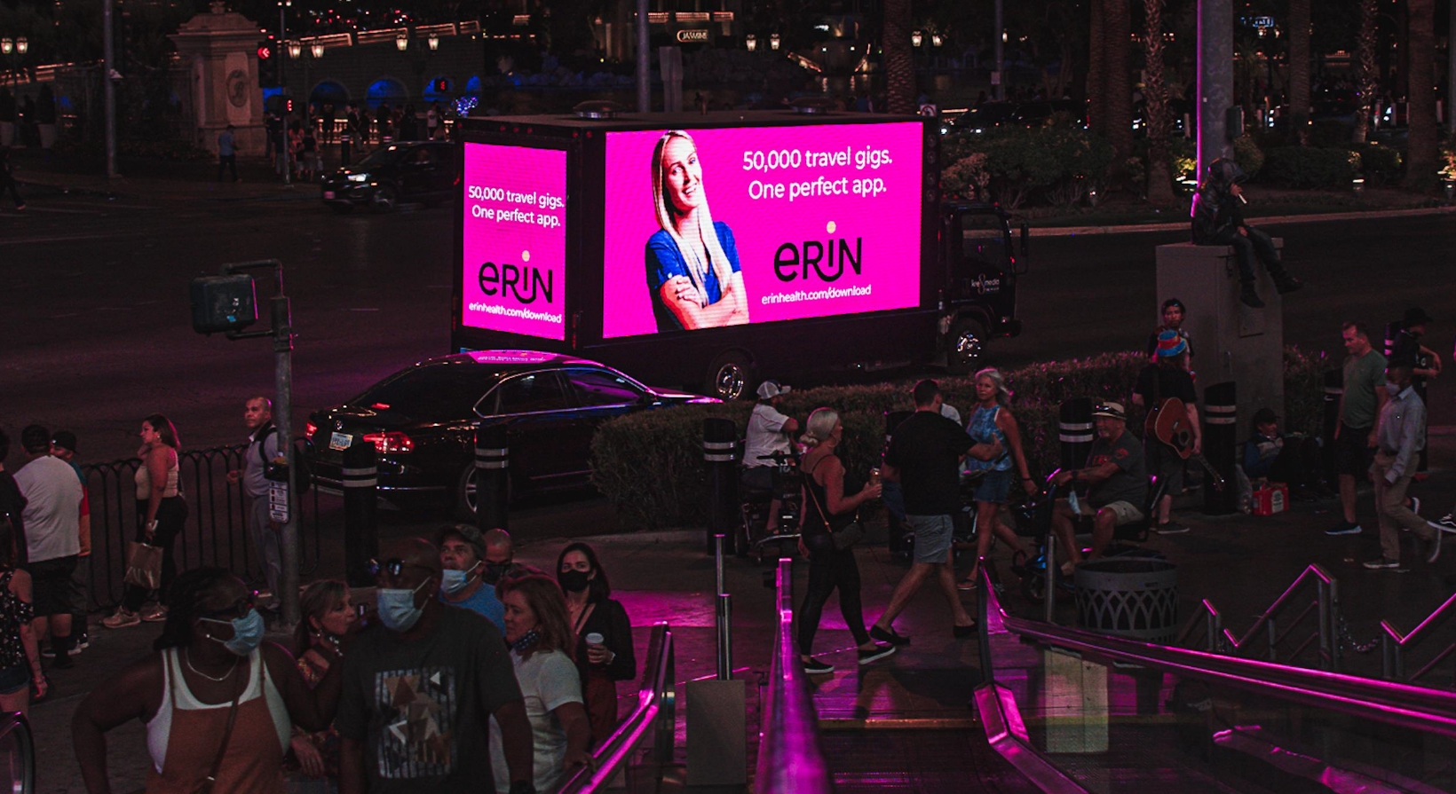 Crowd on a busy street at night with neon lights. A truck with a large digital billboard displays an advertisement for a travel app. People are talking, some wearing masks, and cars are passing by. Stairs lead down to street level.