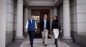 Three people walking outside a building with large pillars. They are engaged in conversation, with two men in business casual attire and a woman in a black top and white skirt. The entrance features a wooden door with glass panels.