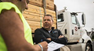 A person in a black jacket stands with arms crossed, talking to another person in a yellow shirt holding a pen and clipboard. A truck loaded with wooden planks is in the background.