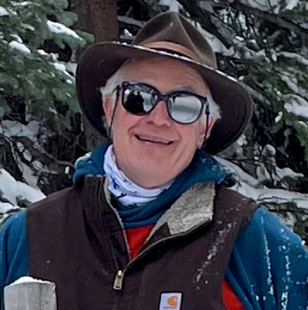 A person wearing a brown hat, dark sunglasses, and winter clothing stands outdoors in a snowy forest. They are smiling and have a blue scarf around their neck.