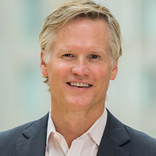 Smiling person with short, light hair wearing a suit and white shirt, standing against a blurred indoor background.
