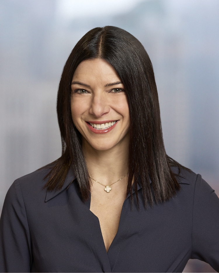 A woman with long, dark hair is smiling and wearing a dark blouse and a delicate necklace. She stands against a blurred background, creating a professional and approachable appearance.