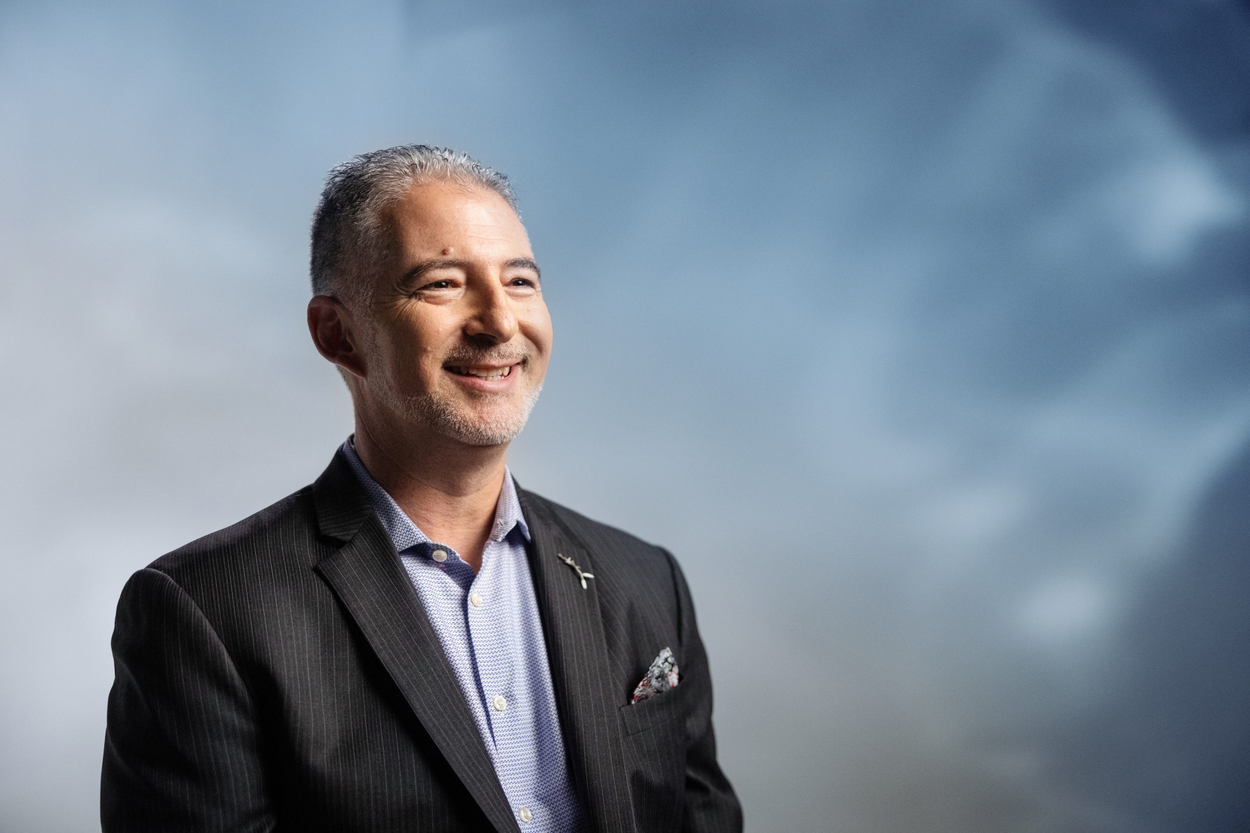 A man in a dark suit and light shirt, smiling against a blurred blue-gray background.