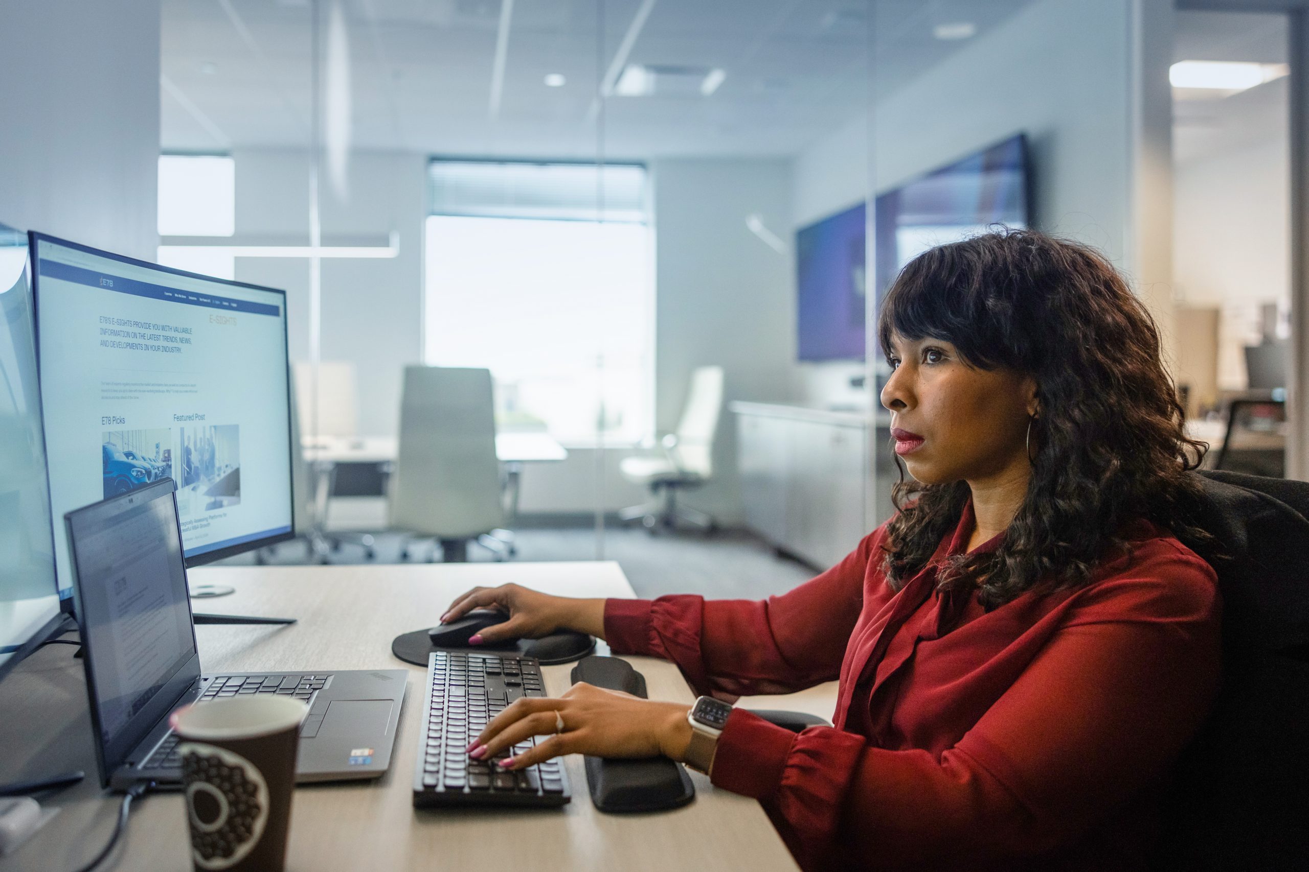 A woman in a red blouse works on a computer in a modern office. She is sitting at a desk with a laptop and a large monitor displaying a webpage. A coffee cup is on the desk, and bright daylight filters through a window.