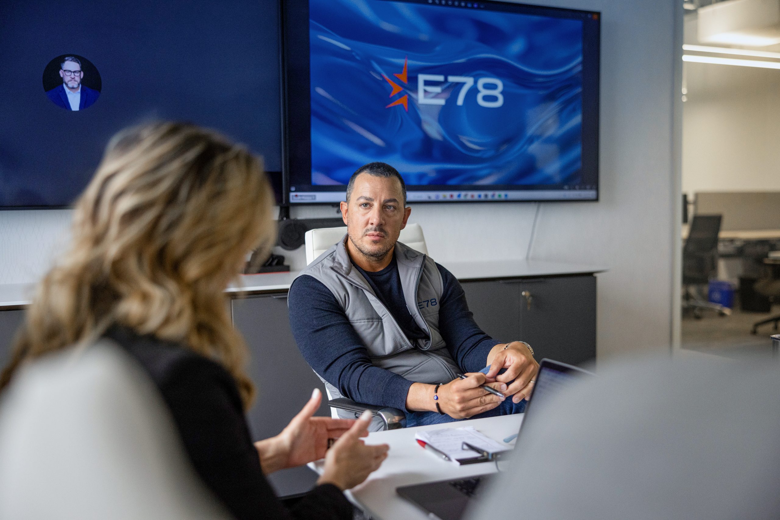 A man is sitting in an office meeting room with E78 displayed on a screen behind him. He is holding a pen and listening to a woman across the table. Another screen shows a participant in a video call.
