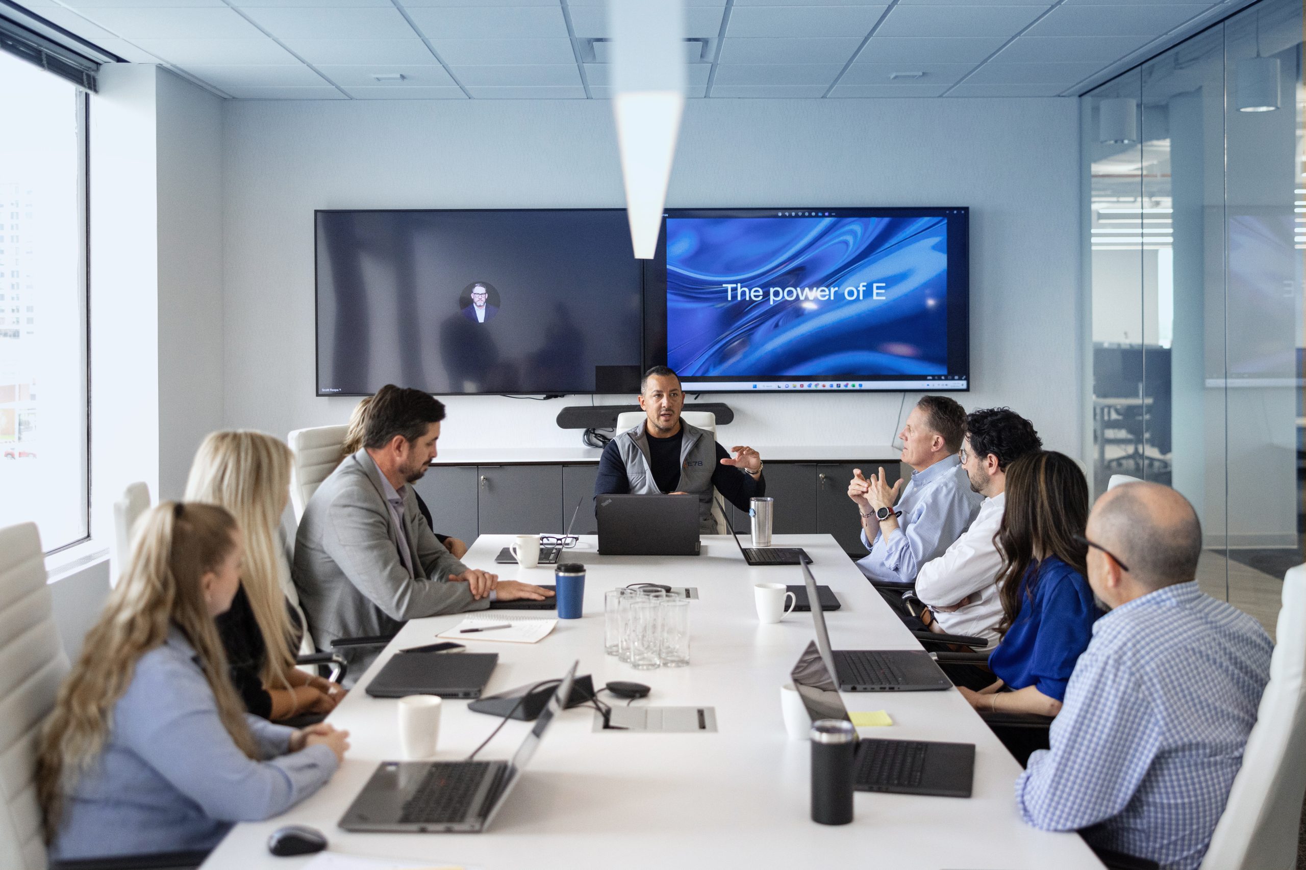 A man presents at a conference table surrounded by colleagues in a modern office. Two screens are on the wall, one with a blue graphic that reads The power of E. Laptops and notebooks are on the table.
