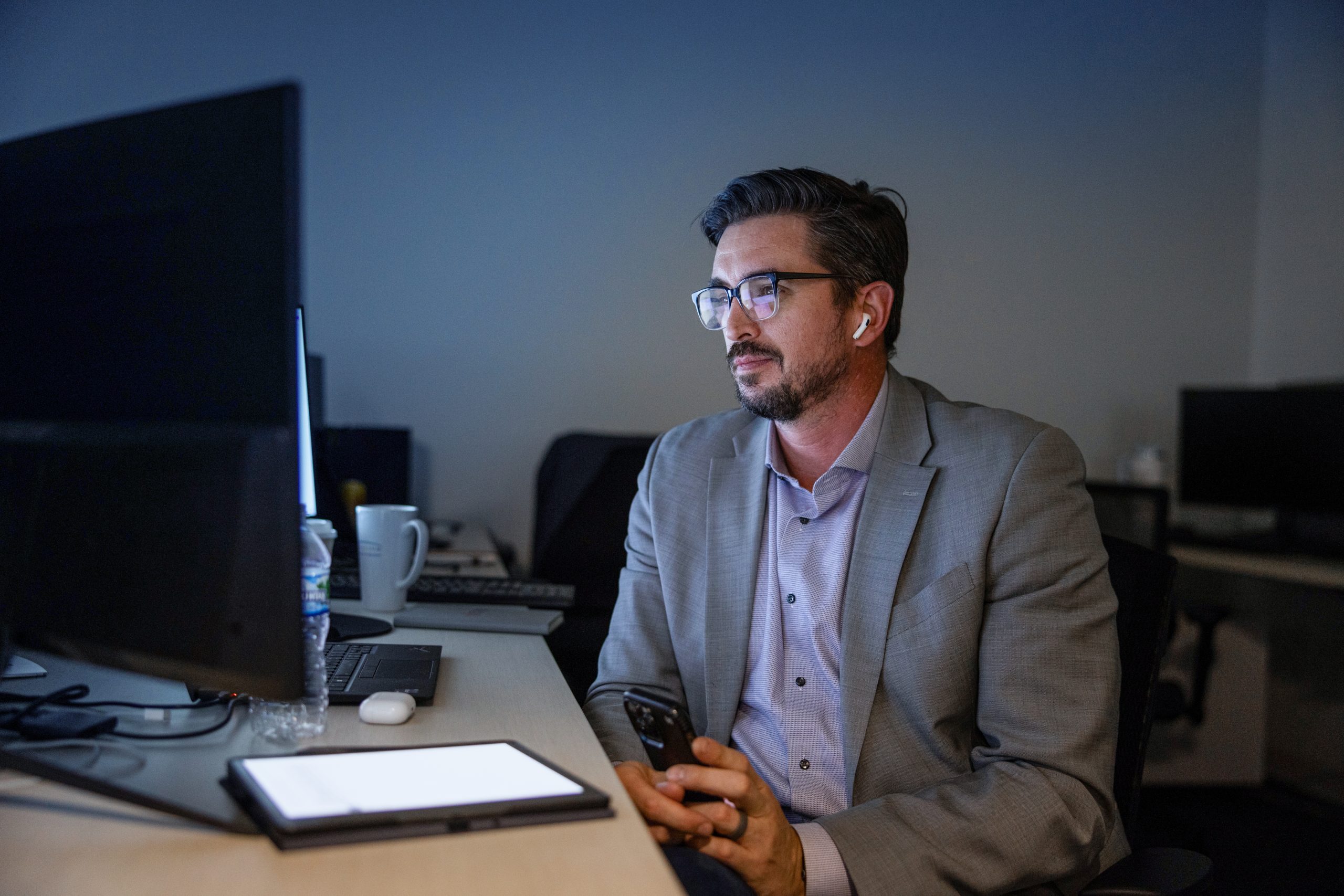 A man in a suit and glasses sits at a desk in a dimly lit office, holding a smartphone and wearing earbuds. He is looking at a computer screen, with various office items like a coffee mug and a water bottle nearby.