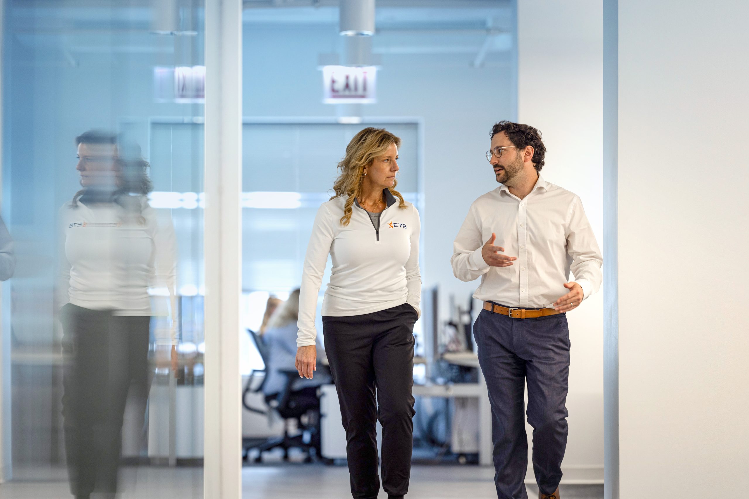 A woman and a man are walking and talking down a hallway in a modern office. They both wear business casual attire. The hallway has glass walls, and there are blurred figures and office equipment in the background.