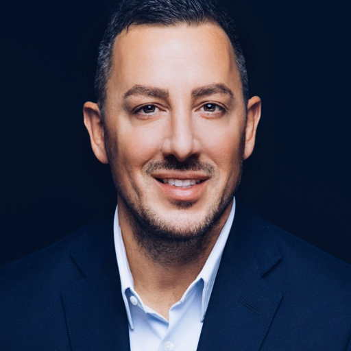 A person with short dark hair and facial hair is smiling, wearing a dark blue suit jacket over a white shirt. The background is a dark, solid color.