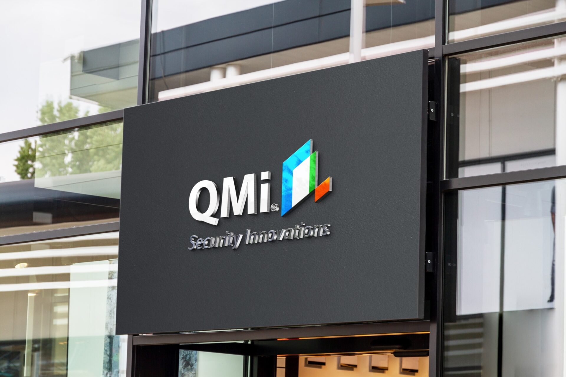 Signboard displaying the QMi Security Innovations logo with colorful geometric shapes on a dark background, mounted on a modern glass building.