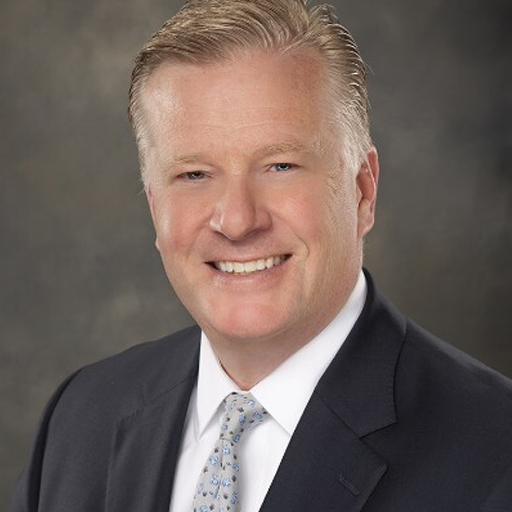 A smiling person with short blonde hair wearing a dark suit, white shirt, and a light patterned tie, set against a dark, textured background.