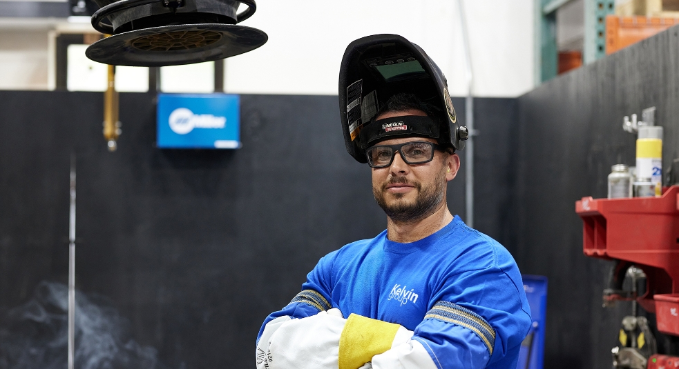 A person wearing a welding helmet and safety glasses stands confidently with arms crossed. They are dressed in a blue shirt and white protective gear, surrounded by tools and welding equipment in a workshop.