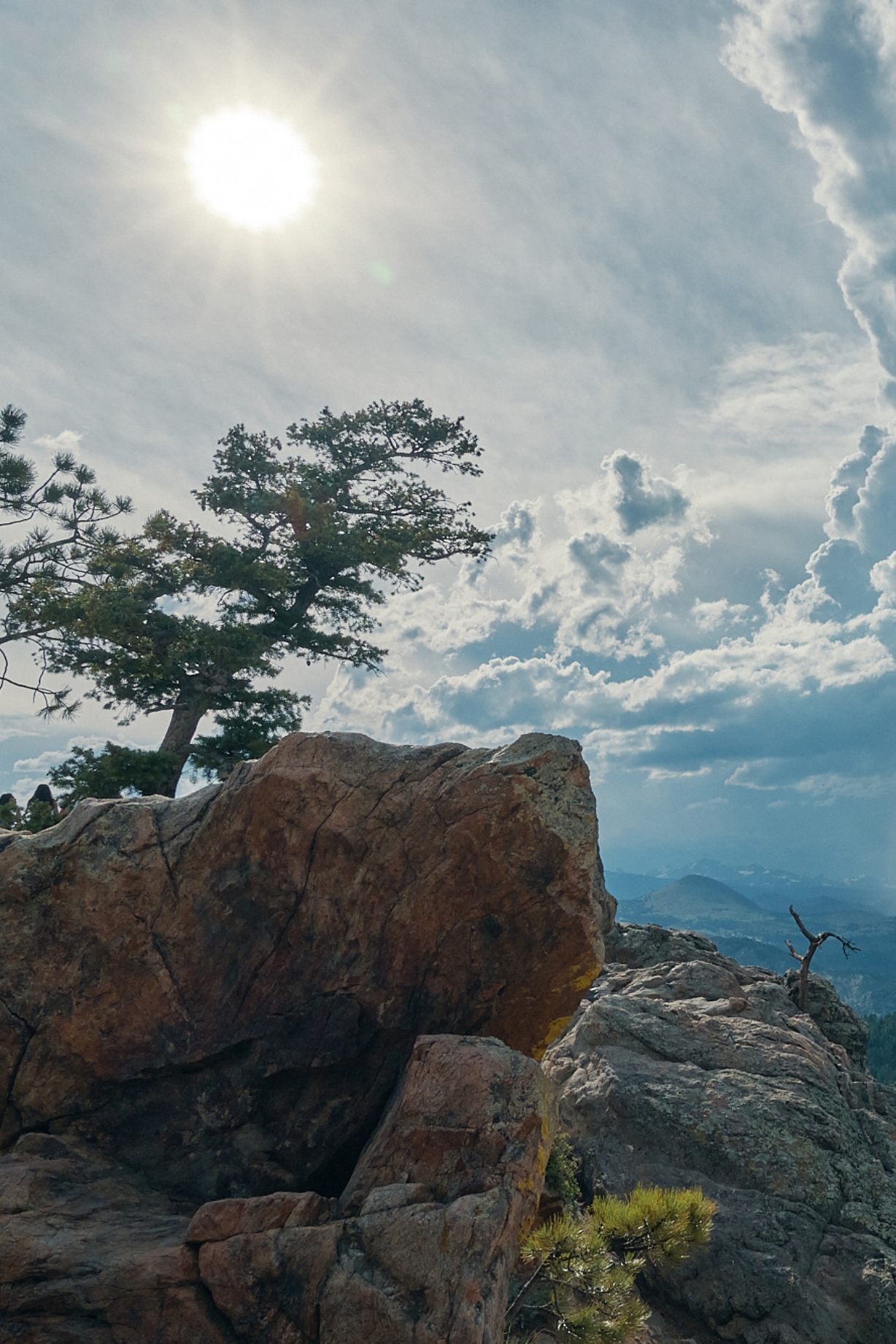 Rocky landscape with a large, sunlit boulder in the foreground, accompanied by a silhouetted tree. The bright sun shines through a partly cloudy sky, casting shadows over distant mountains.