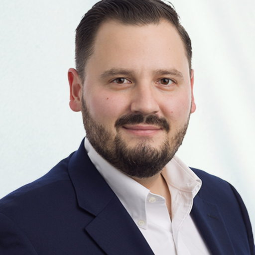 A man with short dark hair and a beard is smiling slightly. He is wearing a white shirt and a dark blazer. The background is a plain light color.