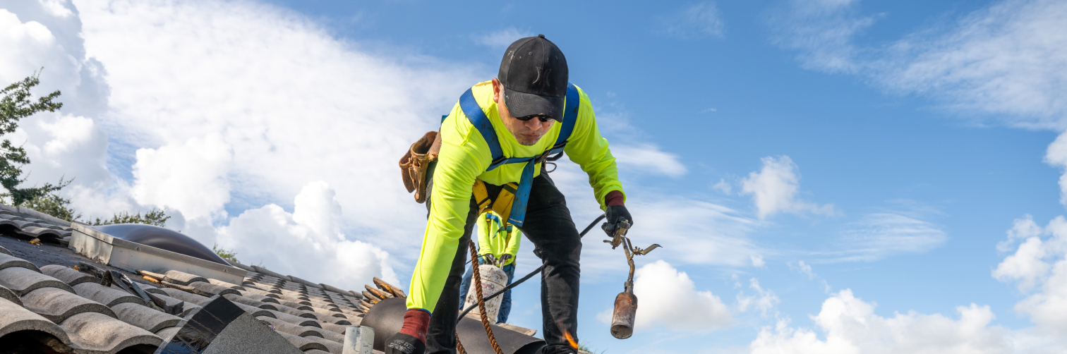 A worker in a bright yellow safety jacket and black cap crouches on a tiled roof under a blue sky. Wearing a safety harness and holding a tool, they are focused on their task, embodying the meticulous attention to detail that private equity firms seek when enhancing portco branding.