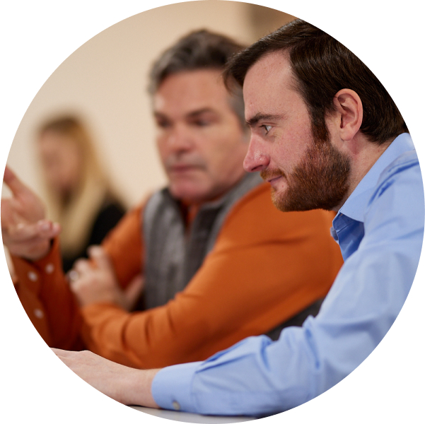 Two men are seated and engaged in a conversation. One, in an orange shirt, is passionately discussing brand strategy, while the other, wearing blue, is attentively focusing on the creative ideas being shared. The background remains blurred.