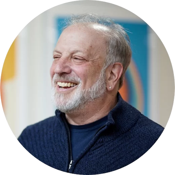 A smiling older man with a white beard and short hair wears a navy sweater over a dark shirt, exuding thought leadership. An earring graces his left ear, set against a colorful, blurred background that enhances his charismatic presence.