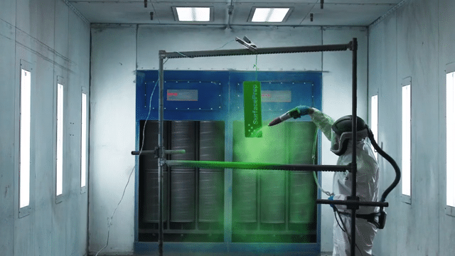 A PE & Portco Expert in protective gear is spray painting a sign with green paint in an industrial booth. The booths blue and gray interior, illuminated by overhead lights, provides the perfect backdrop as the sign hangs vertically for precise application.