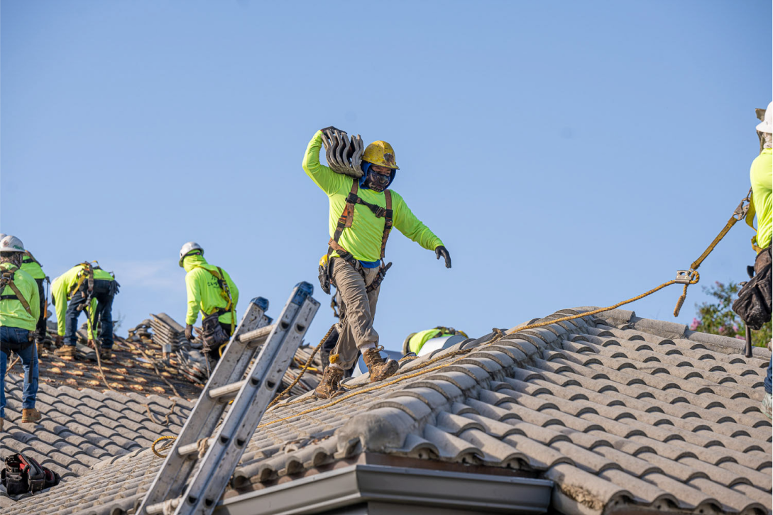 Roofers wearing safety gear work on a tile roof under a clear blue sky. Some carry tiles, while others adjust ropes and equipment. A ladder leans against the roofs edge.