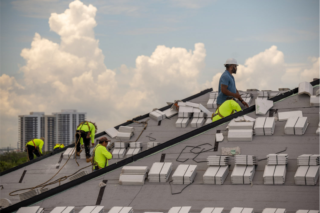Workers in safety gear install roofing tiles on a sloped roof under a partly cloudy sky. A city skyline with tall buildings is visible in the background.