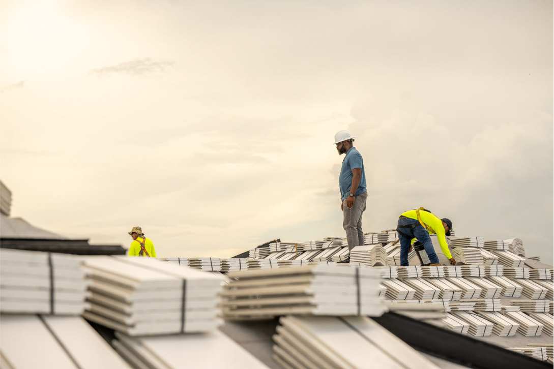 Construction workers in safety gear stand and work on a large stack of drywall panels under a cloudy sky. The foreground shows neatly piled panels, while the background features two workers wearing helmets and reflective vests.