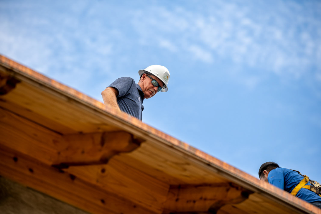 A construction worker in a white hard hat is standing on a wooden roof, looking down. The sky is partly cloudy. Another worker in a blue shirt is partially visible, working on the roof.