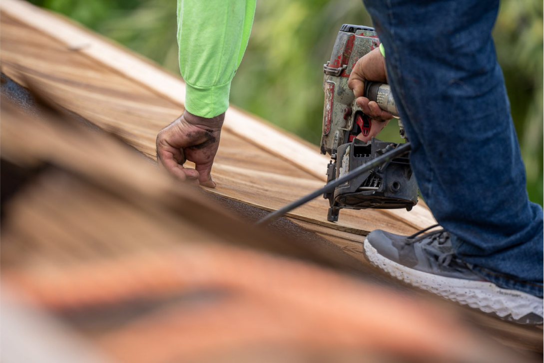 A person in a green shirt and jeans crouches on a roof, using a nail gun. They are securing wooden shingles with one hand while holding the tool with the other. The angle shows a close-up of the action, with blurred shingles in the foreground.