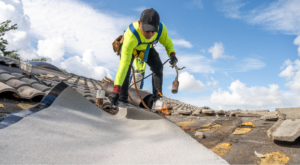A construction worker in a neon yellow jacket and safety harness is working on a roof. He is using a tool to apply roofing material. The sky is bright with scattered clouds. Broken roof tiles are visible nearby.