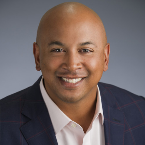 A person with a shaved head is smiling at the camera, wearing a dark blazer and light-colored collared shirt. The background is a plain gray gradient.