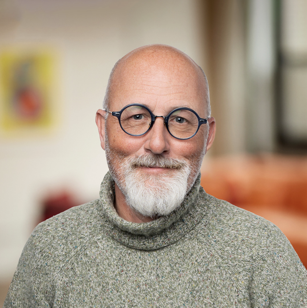 A person with a bald head, a gray beard, and round glasses is smiling. They are wearing a gray turtleneck sweater. The background is blurred, showing soft colors and a framed picture.