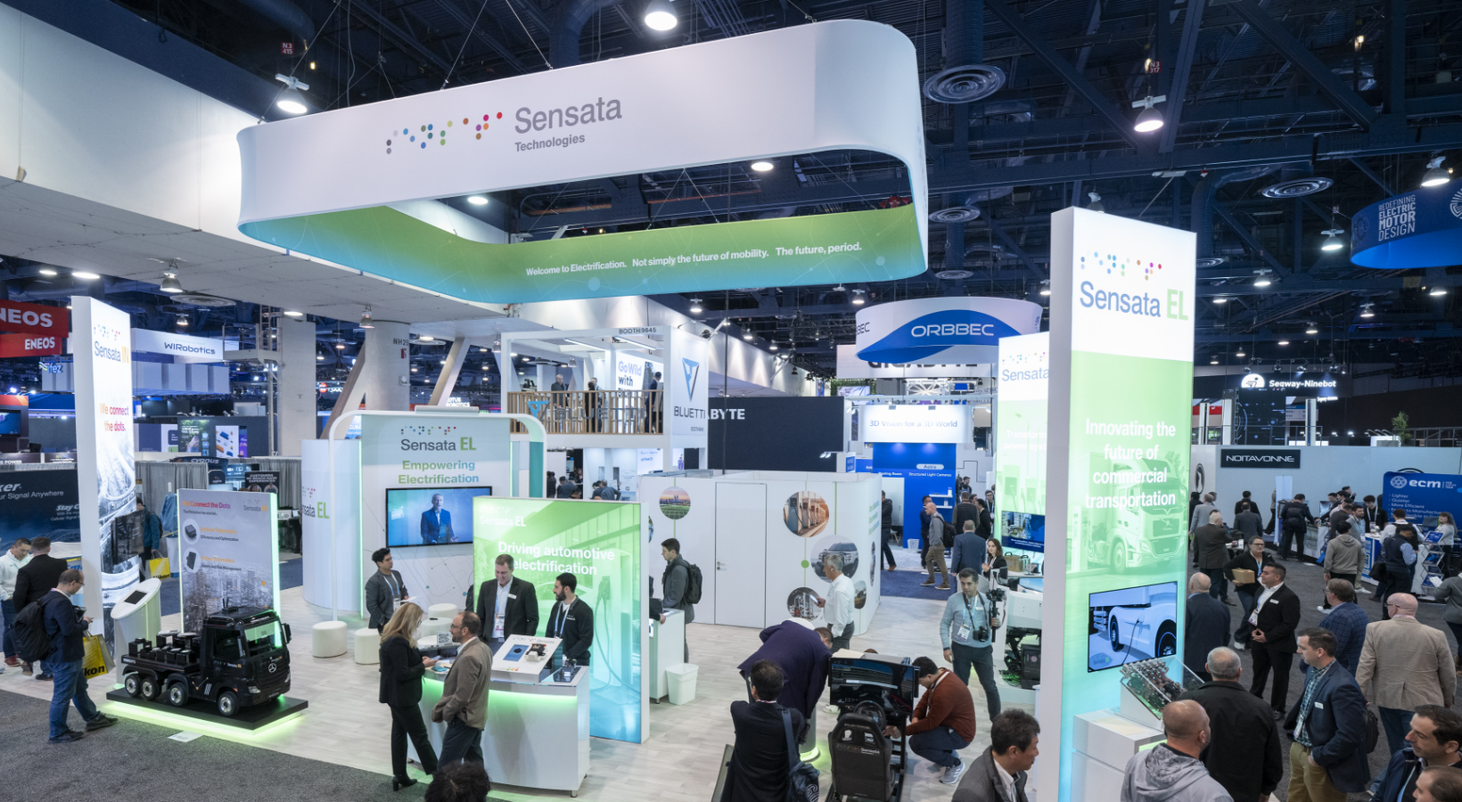 A busy trade show booth with Sensata Technologies branding. Visitors explore displays featuring technology and electrification solutions. The booth is brightly lit, with multiple informational panels and small tech exhibits.