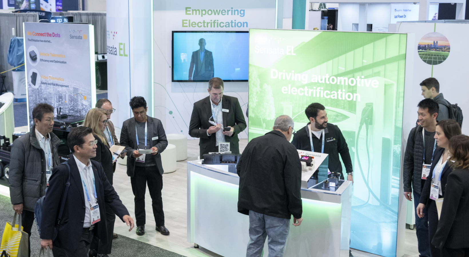 People gather around a booth at a technology expo. A large green sign reads Driving automotive electrification. Attendees are engaged with displays, while others walk past or converse. Posters in the background highlight related technology themes.