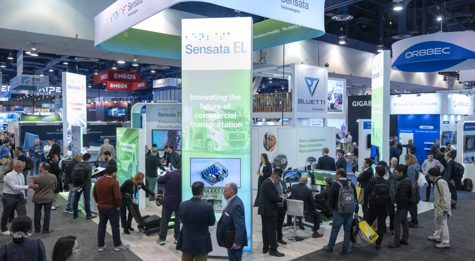 A busy trade show booth featuring a prominent Sensata EL display with the tagline Innovating the future of commercial transportation. Visitors are engaging with various exhibits and screens. Other company booths and attendees are visible around the area.