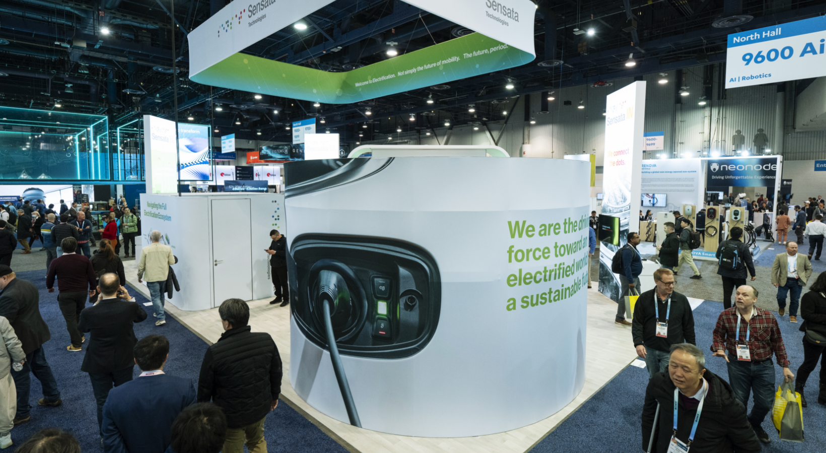 A busy trade show floor with people walking around exhibition booths. A large, prominent booth features a display of an electric vehicle charging port. Overhead banners are visible, and various signs and products are showcased throughout the space.
