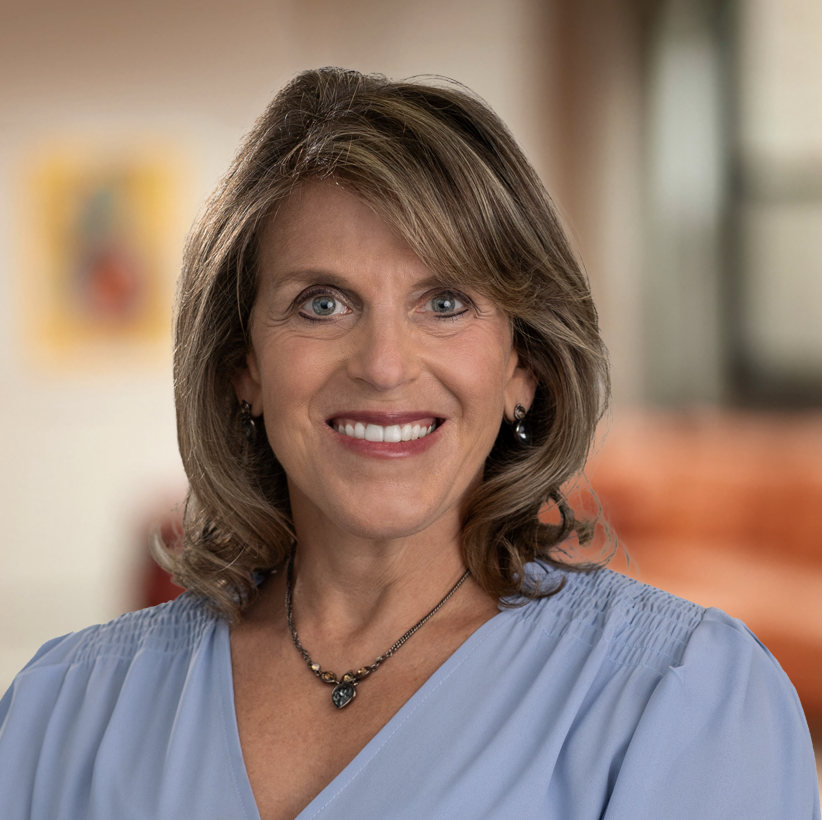 A person with shoulder-length hair smiles at the camera, wearing a light blue blouse and a necklace. The background is a softly focused room with warm colors.