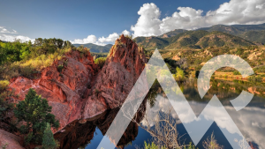 Scenic view of a rocky, red sandstone formation with lush green vegetation around it. It is reflected in a calm lake. Mountains and fluffy clouds are visible in the clear blue sky. A transparent logo is overlaid on the image.
