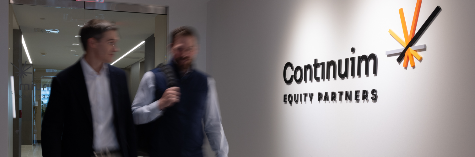 Two men walk past a wall displaying the Continuum Equity Partners logo—its stylized lines in orange, white, and black reflecting both private equity strength and distinctive portco branding.