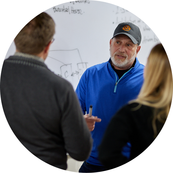 A man in a blue pullover and gray cap discusses private equity with two people in front of a whiteboard filled with handwritten diagrams and text. He holds a marker, actively engaging in the conversation.