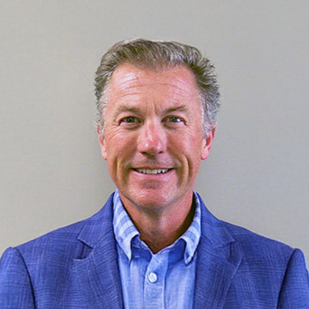 A middle-aged man with short gray hair is smiling at the camera. He is wearing a light blue collared shirt and a blue blazer, standing in front of a plain light-colored background.