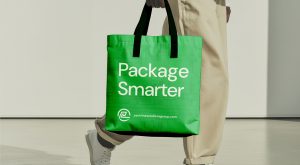 Person holding a bright green tote bag with black handles. The bag features the text Package Smarter and the website packlinesolutionsgroup.com in white letters. The person is dressed in light-colored pants.