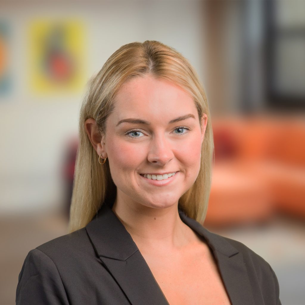 A young woman with straight blonde hair, wearing a black blazer and gold hoop earrings, smiles at the camera. The background is an office or living room, softly blurred with warm tones.